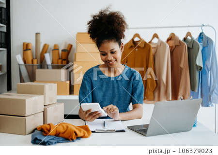 Young African woman running online store Startup small business SME, using smartphone or tablet taking receive and checking online purchase shopping order in office 106379023