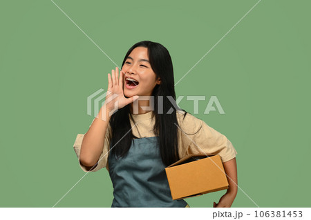 Teenage Asian girl holding parcel box and shouting with hands on mouth, standing against green background Teenage Asian girl holding parcel box and shouting with hands on mouth, standing against green background 106381453