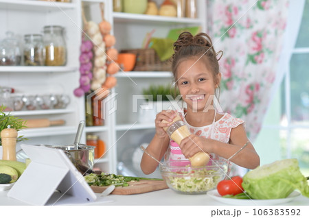 little girl preparing fresh salad on kitchen table with tablet at home 106383592