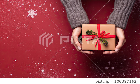 Festive Joy Female Hands Holding Christmas Gift Box with Evergreen Decor on Snowy Red Background. created with Generative AI 106387162