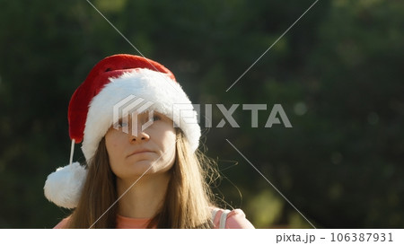 A young woman in a red Christmas hat walks against the background of a green forest. A warm Christmas. 106387931