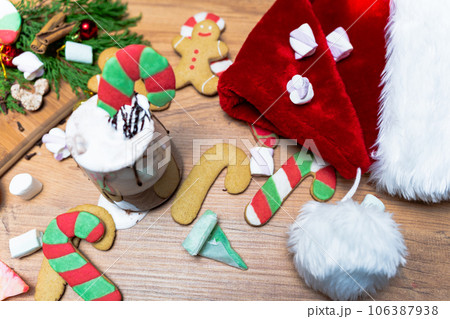 Gingerbread candy cane in a cup of hot chocolate with whipped cream, Christmas interior. Santa's hat on the table, top view. Marshmallows scattered everywhere. 106387938