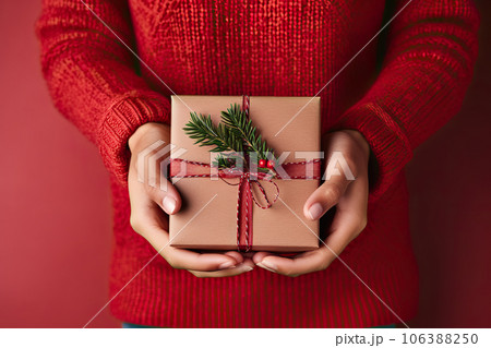 Festive Gift Giving Female's Hands Holding Christmas Gift Box with Evergreen Branch on Red Background. created with Generative AI Festive Gift Giving Female's Hands Holding Christmas Gift Box with Evergreen Branch on Red Background. created with Generative AI 106388250