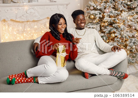 Happy black couple holding gift and sitting on a coach near Christmas tree. Man amd woman celebrating winter holidays at home. Brunette woman wearing a red sweater. 106389514