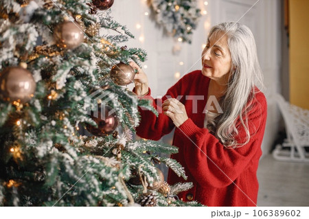 Charming old female is preparing for New Year. Senior woman standing near christmas tree. Woman has gray hair and wearing red sweater. 106389602