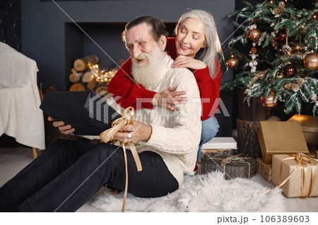 Senior man celebrating christmas with his wife near beautiful christmas tree. Old bearded man sitting on a floor near his wife with grey hair and holding a gifts. Man wearing white sweater and woman 106389603