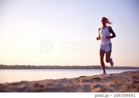 Full length portrait of young healthy woman in sunglasses, training, running at beach near river on sunrise. Morning fitness exercise in motion 106389891