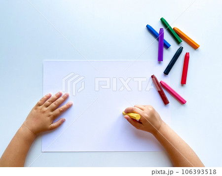 Childrens hands draw with colored wax crayons on a white sheet of paper. Top view of a blank sheet. Childrens hands draw with colored wax crayons on a white sheet of paper. Top view of a blank sheet. 106393518