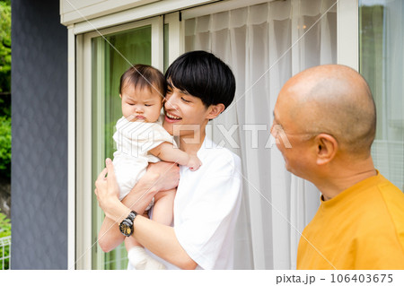 屋外の家の前で0歳児の赤ちゃんを抱っこする父親とそれを見守る祖父 屋外の家の前で0歳児の赤ちゃんを抱っこする父親とそれを見守る祖父 106403675