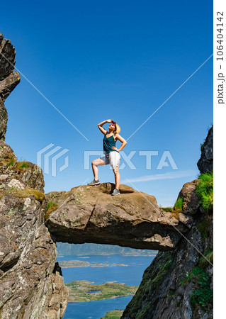 Brave traveler woman standing on hanging stone between rocks. Djevelporten in Norway Lofoten islands Brave traveler woman standing on hanging stone between rocks. Djevelporten in Norway Lofoten islands 106404142