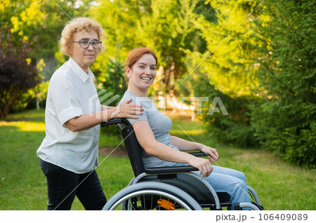 An elderly woman is carrying an adult daughter sitting in a wheelchair. Walk outdoors.  106409089