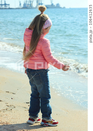 Little girl with long blond hair pink coat plays on the beach with sand shells stones looks at the sea waves sunny day. 106412635