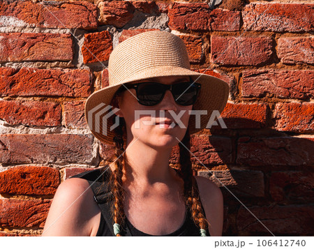 Young woman in straw hat portrait on red brick wall urban background Pretty millennial girl walks on summer city street Young woman in straw hat portrait on red brick wall urban background Pretty millennial girl walks on summer city street 106412740