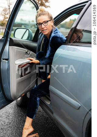 Blonde woman in glasses look back over her shoulder as she exits parked blue car in which she drove. 106414369