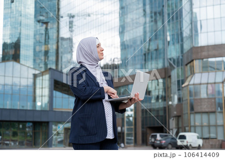Thoughtful business woman in hijab and suit is holding a laptop outdoors. 106414409