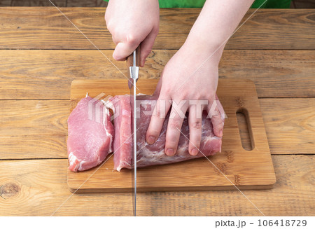 A young woman cuts raw pork meat on a cutting board with a knife. 106418729