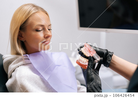 Close-up cropped shot of unrecognizable female dentist in protective mask and gloves showing jaw model with braces to young woman patient in dental office. Concept of stomatology, orthodontics. 106426454