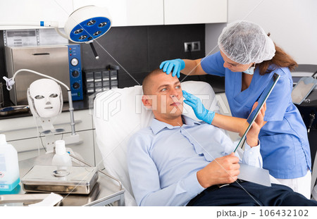 Female cosmetician explaining man hardware facial procedure in cosmetological office Female cosmetician explaining man hardware facial procedure in cosmetological office 106432102