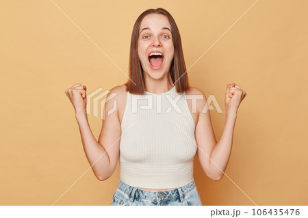 Amazed brown haired young woman wearing casual clothing standing isolated over beige background screaming loud with widely open mouth clenched fists Amazed brown haired young woman wearing casual clothing standing isolated over beige background screaming loud with widely open mouth clenched fists 106435476