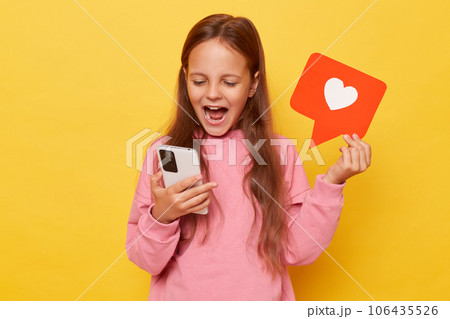 Clicking the like button on a blog post. Recommending viral content to followers. Amazed overjoyed little girl holding blogger heart like icon and smartphone isolated over yellow background 106435526