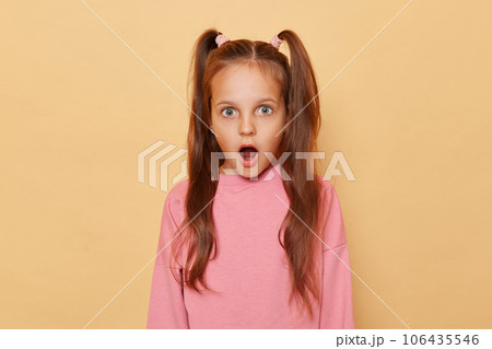 Shocked surprised little girl with ponytails wearing pink sweatshirt isolated over beige background looking at something astonished with widely open mouth and big eyes. 106435546