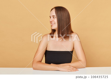 Cheerful smiling brown haired woman wearing black top posing isolated over beige background looking away at copy space for advertisement mockup for promotional text. Cheerful smiling brown haired woman wearing black top posing isolated over beige background looking away at copy space for advertisement mockup for promotional text. 106435725