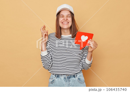 Hopeful woman wearing striped shirt and baseball cap holding like blogger icon standing isolated over beige background crossing fingers dreaming to get a lot of likes on social networks Hopeful woman wearing striped shirt and baseball cap holding like blogger icon standing isolated over beige background crossing fingers dreaming to get a lot of likes on social networks 106435740