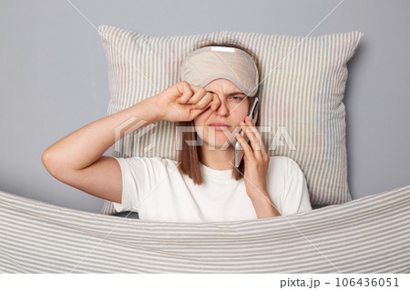 Sleepy young woman wearing white T-shirt and sleep eye mask lies under blanket in bedroom isolated on gray background talking on smartphone feeling sleepy rubbing her eye. 106436051