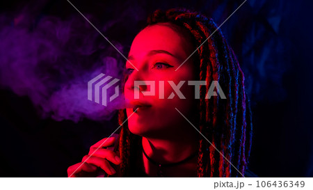 Caucasian girl with dreadlocks smokes a vape in red blue light.  106436349