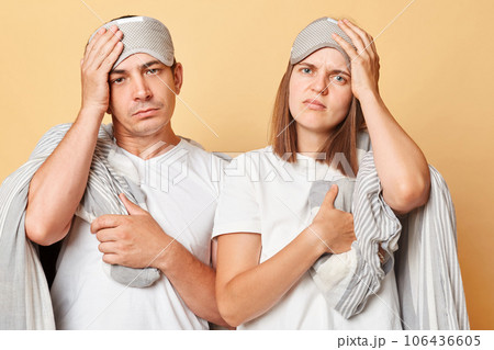 Oversleep couple man and woman in sleep eye mask wrapped in blanket isolated over beige background waking up early having headache need rest. Oversleep couple man and woman in sleep eye mask wrapped in blanket isolated over beige background waking up early having headache need rest. 106436605