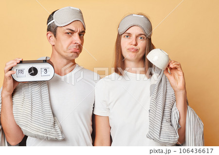 Displeased couple man and woman in sleep eye mask wrapped in blanket holding cup and alarm isolated over beige background waking up early showing empty mug needs coffee. 106436617