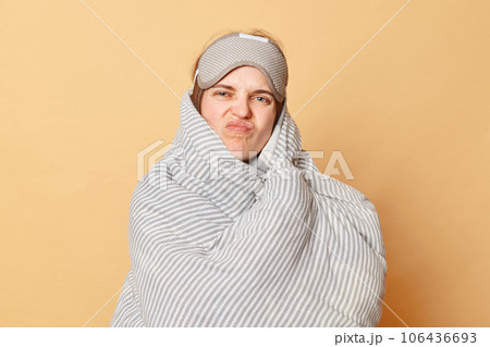Displeased unhappy woman wrapped blanket and wearing sleeping eye mask isolated over beige background does not want to wake up early grimacing frowning face. Displeased unhappy woman wrapped blanket and wearing sleeping eye mask isolated over beige background does not want to wake up early grimacing frowning face. 106436693