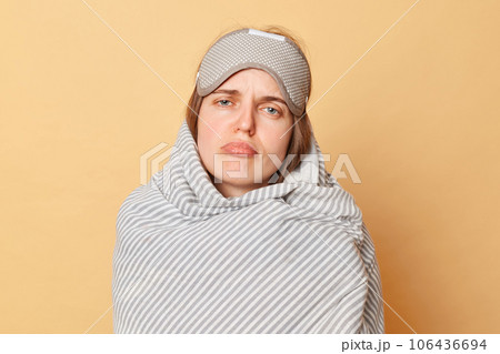 Unhappy sad woman wrapped blanket and wearing sleeping eye mask isolated over beige background looking at cmaera with pout lips lazy to wake up early. Unhappy sad woman wrapped blanket and wearing sleeping eye mask isolated over beige background looking at cmaera with pout lips lazy to wake up early. 106436694