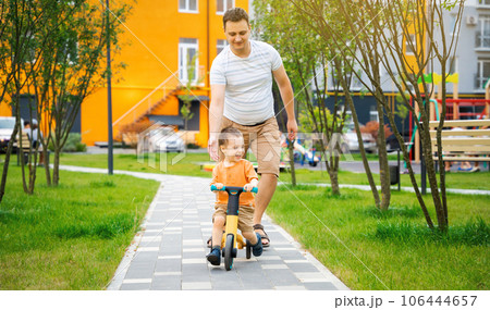 A cute happy toddler boy of two or three years old with dad rides a bicycle or balance bike in a residential complex on a sunny summer day. Happiness spending time with family A cute happy toddler boy of two or three years old with dad rides a bicycle or balance bike in a residential complex on a sunny summer day. Happiness spending time with family 106444657