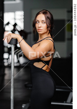 Female bodybuilder with long hair posing in gym with sports equipment. Strong woman with muscular body leaning on barbells in gym in dark atmosphere, looking at camera. 106444892