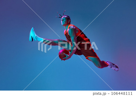 Young sportive athlete, basketball player in motion jumping with ball against blue studio background in neon light 106448111