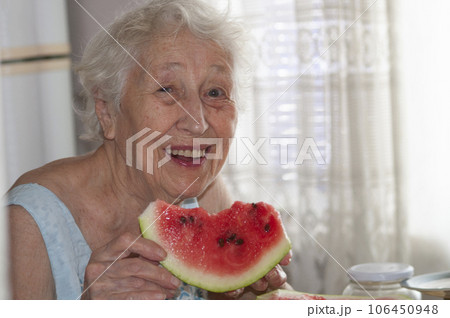 Smiling old senior woman holding a slice of fresh watermelon looking at camera at home background - healthy eating lifestyle concept 106450948