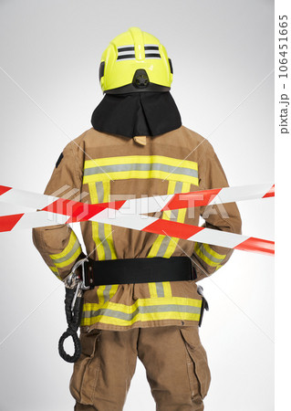 Strong unrecognizable firefighter in helmet standing behind red and white caution tape. Back view of male fireman working in closed by caution stripe area, on white background. Workplace concept. Strong unrecognizable firefighter in helmet standing behind red and white caution tape. Back view of male fireman working in closed by caution stripe area, on white background. Workplace concept. 106451665