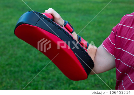 Man's hand holds a red makiwara for punches and kicks against the background of grass in a street training for practicing punches in a fight 106455314