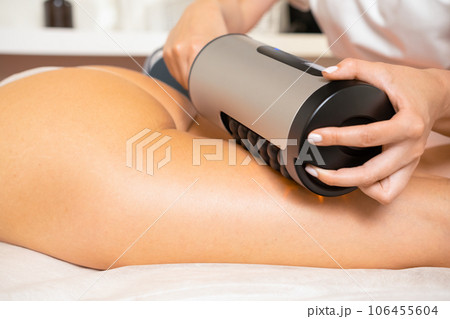 Electric massage device for figure correction in clinic room. Beautician doing massage of clients hip thigh and legs with vibrational therapy apparatus Electric massage device for figure correction in clinic room. Beautician doing massage of clients hip thigh and legs with vibrational therapy apparatus 106455604