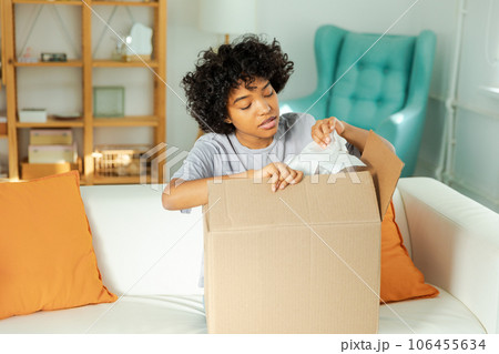 African girl unpacking delivery looking in box. Happy woman opening carton box. Female getting parcel looking at delivered goods items. Satisfied client positive feedback quick express deliver African girl unpacking delivery looking in box. Happy woman opening carton box. Female getting parcel looking at delivered goods items. Satisfied client positive feedback quick express deliver 106455634