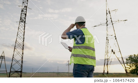 Electrician conducts diagnostic of power transmission lines with tablet in field. Engineer with tablet works at electrical power plant in field. Skilled engineer controls work of electric power lines 106457420