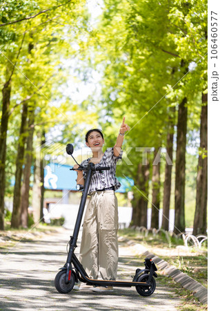 爽やかな木漏れ日の中を電動キックボードで散策する若くてかわいい女性｜電動キックボードイメージ 106460577