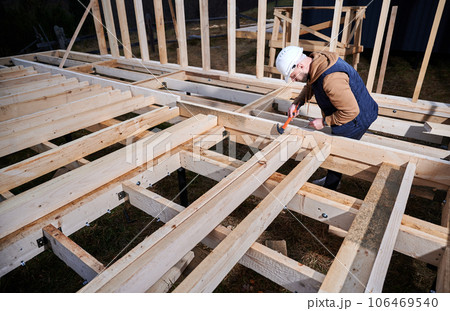 Man worker building wooden frame house on pile foundation. Carpenter hammering nail into wooden joist, using hammer. Carpentry concept. Man worker building wooden frame house on pile foundation. Carpenter hammering nail into wooden joist, using hammer. Carpentry concept. 106469540