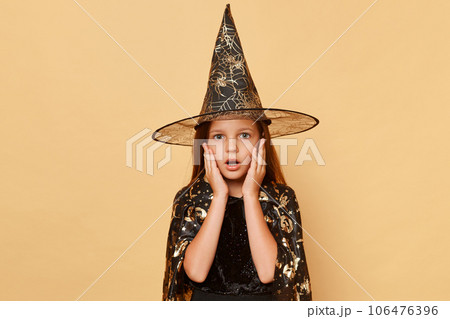 Magical wizard and sorcerer. Astonished little girl wearing witch costume and carnival cone hat isolated over beige background keeps hands on cheeks looking with big eyes at something scary. Magical wizard and sorcerer. Astonished little girl wearing witch costume and carnival cone hat isolated over beige background keeps hands on cheeks looking with big eyes at something scary. 106476396