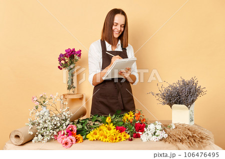 Placed order at floral store. Florist arranged beautiful bouquet. Brown haired cute positive woman wearing green overall and making notes receiving order in florist shop with various flowers Placed order at floral store. Florist arranged beautiful bouquet. Brown haired cute positive woman wearing green overall and making notes receiving order in florist shop with various flowers 106476495