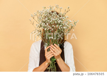Unknown unrecognizable young caucasian woman florist wearing brown apron holding flowers isolated over beige background hiding her face behind bouquet Unknown unrecognizable young caucasian woman florist wearing brown apron holding flowers isolated over beige background hiding her face behind bouquet 106476505