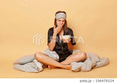 Displeased sad young girl sitting floor with blanket wearing black pajama and sleep eye mask holding plate in hands isolated over beige background felling nausea bed smell of breakfast meal 106476524