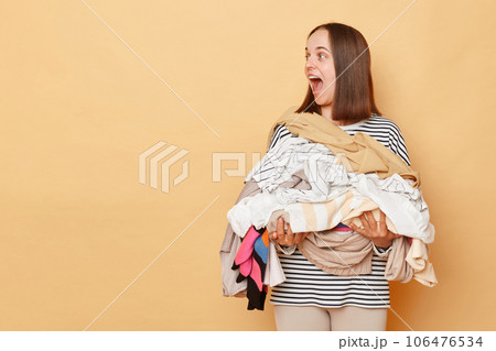 Excited brunette woman holding pile of clothes in hands isolated over beige background looking away at copy space for promotion advertisement area screaming with happiness. Excited brunette woman holding pile of clothes in hands isolated over beige background looking away at copy space for promotion advertisement area screaming with happiness. 106476534