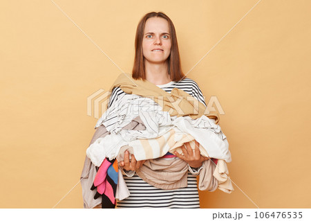 Confused brunette woman holding pile of clothes in hands isolated over beige background looking at camera with confused expression biting her lip. 106476535